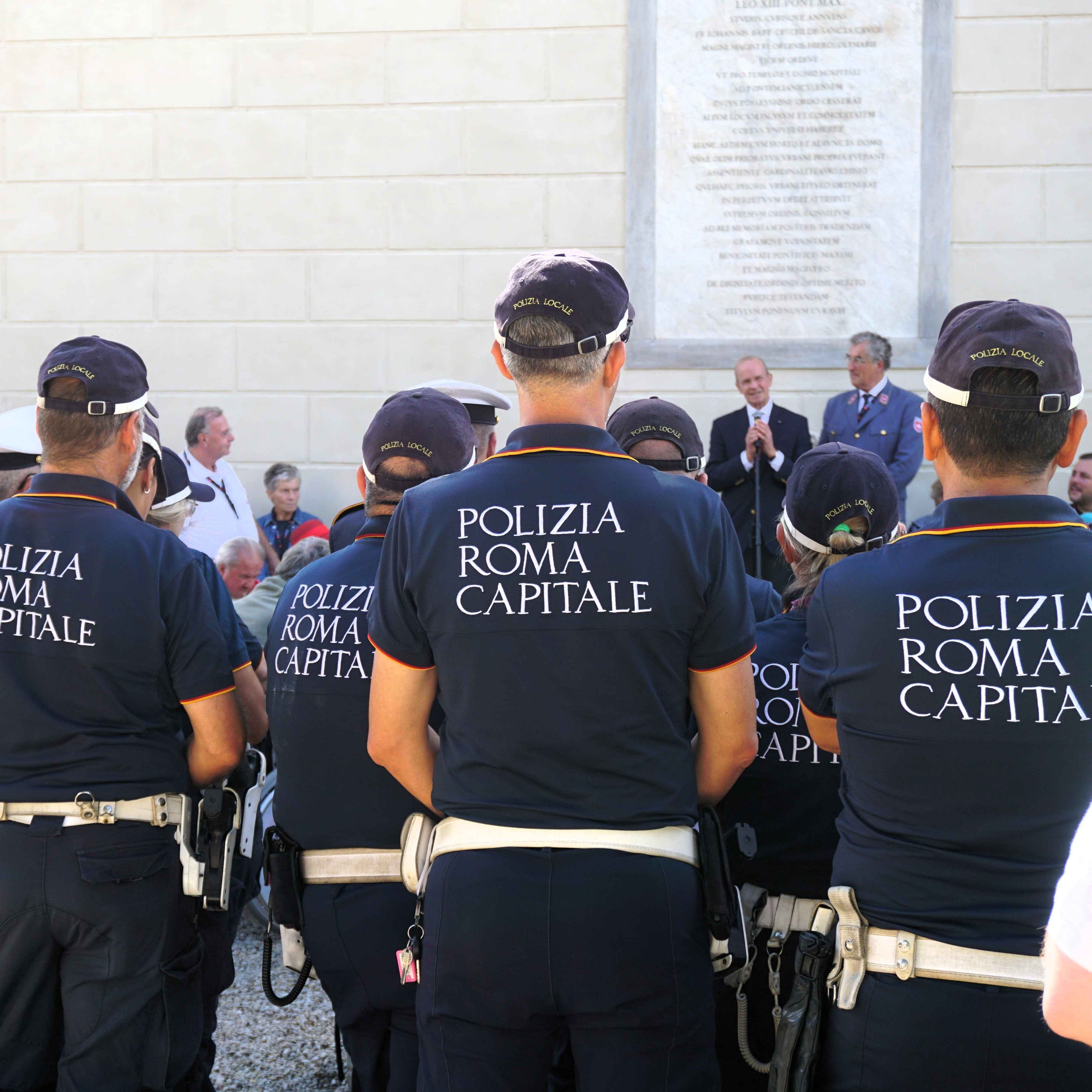 Polizisten der Polizia Roma Capitale in dunkler Uniform mit weißem Gürtel stehen mit dem Rücken zur Kamera vor einer hellen Wand in der Villa Malta in Rom..