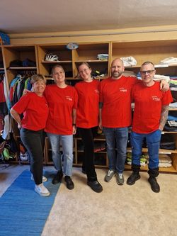 Auf dem Foto sieht man die Mitarbeitenden der Firma Camurus welche am Social Day teilgenommen haben. Sie tragen rote T-Shirts und stehen vor den Regalen der Kleiderkammer.