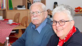 Frau mit grauen kurzen Haaren in Malteser Kleidung und Senior lächelnd in einem Aufenthaltsraum