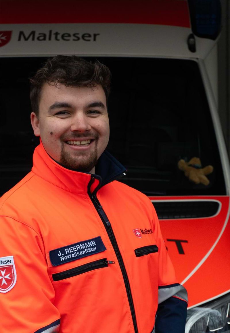 Ein junger Mann in einer leuchtend orangefarbenen Einsatzjacke steht lächelnd vor einem Rettungswagen der Malteser. Auf seiner Einsatzjacke sind ein Namensschild und das Logo der Malteser zu sehen.
