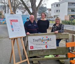 Ein Malteser-Ehrenamtler steht mit zwei Kooperationspartnerinnen im Freien an einem Schild mit dem Titel "Treffpunkt"