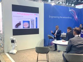Mann bei Präsentation auf Fachmesse, medizinische Inhalte auf Leinwand, Roboter vorne links, Publikum an Tischen.