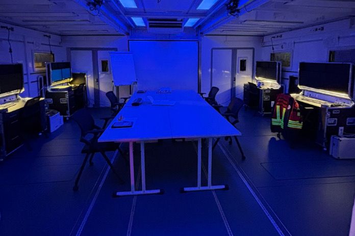 Das Foto zeigt den MedExTainer von innen als einen dunklen Raum, der mit blauem Licht beleuchtet ist. In der Mitte stehen zwei weiße Konferenztische, umgeben von mehreren leeren Bürostühlen. Vorne befindet sich eine große Leinwand oder Projektionsfläche, daneben ein Flipchart. Links und rechts im Raum stehen Arbeitsplätze mit Monitoren, Computerausrüstung und Koffern. Auf der rechten Seite hängt eine Kennzeichnungsweste über einem Stuhl.