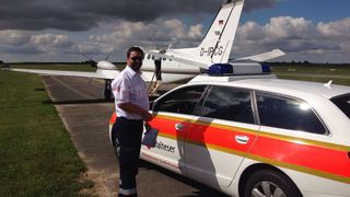 Malteser Mitarbeiter steht neben einem Malteser Wagen vor einem Flugzeug