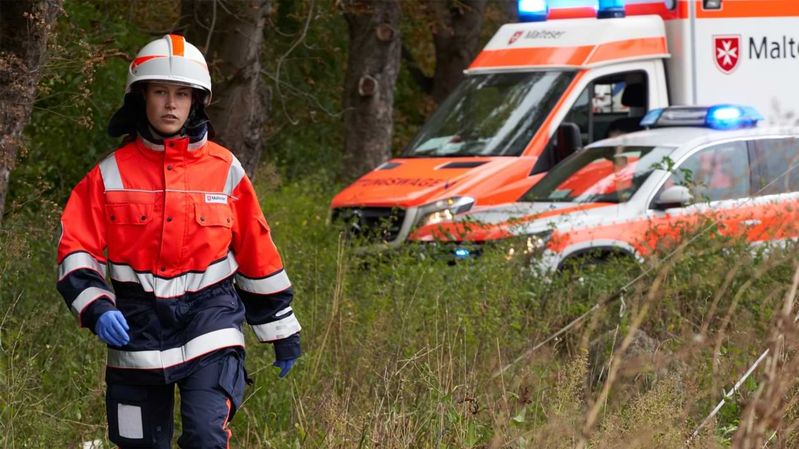 Eine Frau in Einsatzkleidung der Malteser auf einer Lichtung mit Einsatzfahrzeugen im Hintergrund