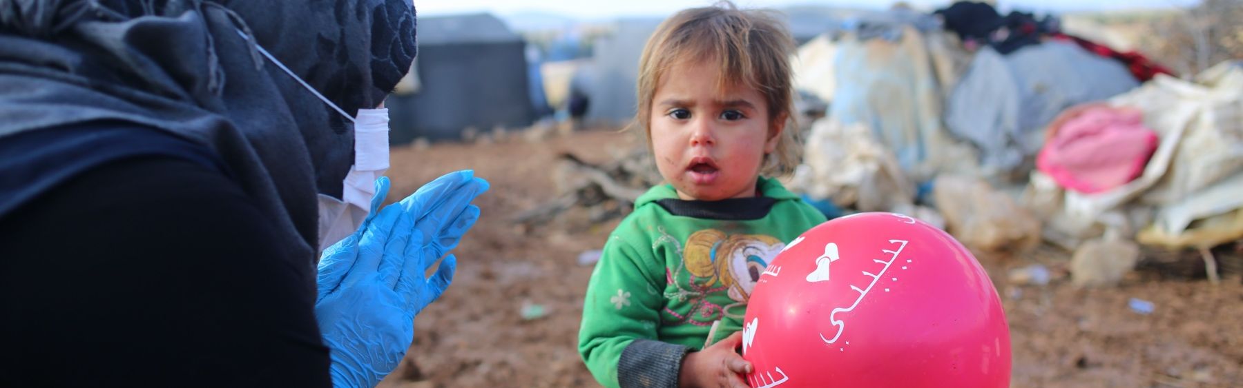 Kind mit einem Luftballon und eine Frau mit Kopftuch und Hygienehandschuhen in einem Flüchtlingscamp.