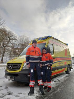 Michelle Richter (rechts) und René Kluge (links) stehen vor einem Rettungswagen der Malteser und lächeln in die Kamera. Die Szenerie ist winterlich.