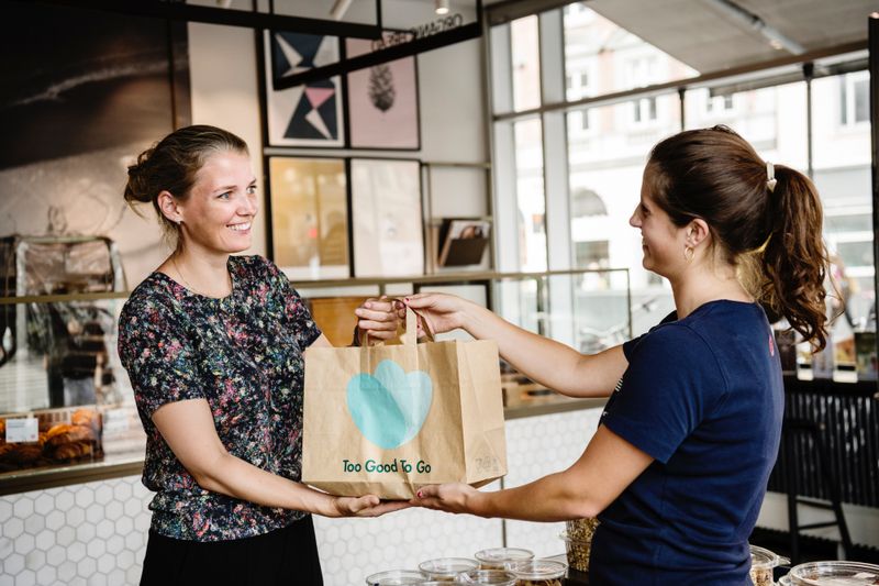 Tag eine Frau überreicht einer anderen Frau eine Papiertüte mit dem Aufdruck „Too Good To Go“