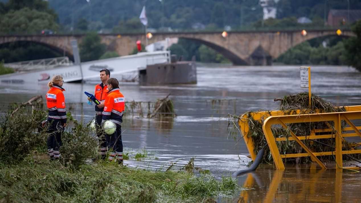 Drei Einsatzkräfte des Katastrophenschutzes stehen an einem überschwemmten Flussbett.
