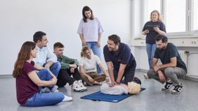 Ein Mann mit Malteser T-Shirt kniet am Boden und zeigt mehren Personen, die um ihn herum stehen oder sitzen, an einer Übungspuppe die Herzdruckmassage. 