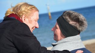 Eine Frau lächelt eine Frau im Rollstuhl an. Beide sind am Meer.