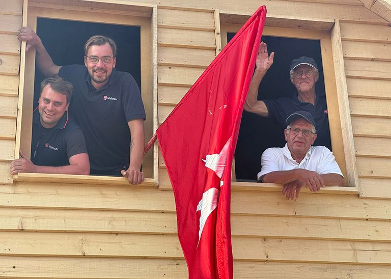 Vier Männer winken aus den Fenstern eines Holzhauses heraus. Am Haus ist eine Malteser Fahne befestigt.