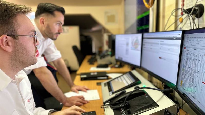 Zwei Männer in einer modernen Funkzentrale mit mehreren Bildschirmen.