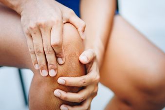 Person hält sich mit beiden Händen schmerzhaft das Knie, sitzend und in sportlicher Klei-dung.
