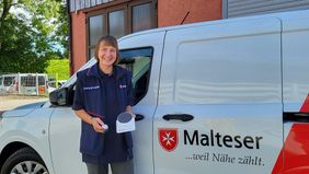 Sabine Meyer von den Rosenheimer Maltesern steht vor einem Malteser Auto und hält ein Hausnotrufgerät in den Händen.