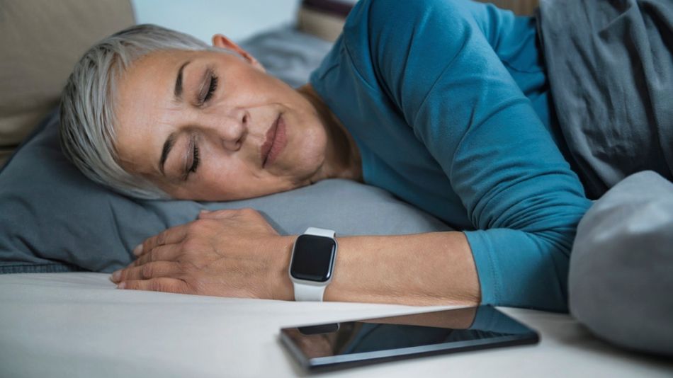 Ältere Frau liegt mit Smartphone und Smartwatch im Bett und schläft.