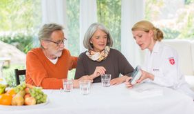 Auf dem Foto sind drei Personen zu sehen, die hinter einem Tisch sitzen. Eine Frau in Malteserkleidung zeigt den beiden anderen Personen, d.h. einem älteren Pärchen, das Hausnotrufgerät. 