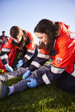 Zwei Sanitäterinnen in Einsatzkleidung kümmern sich um einen Patienten, der auf der Wiese liegt. Sie knien neben ihm. Eine der Sanitäterinnen untersucht sein Schienbein. Die andere öffnet den Sanitätsrucksack, um etwas herauszuholen.