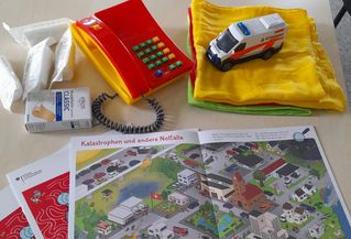 Kinderbuch, das eine Rettungsstelle von oben zeigt, Verbandszeug, ein buntes Spielzeugtelefon und ein Modell eines Rettungswagens.