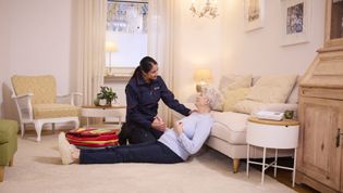Frau mit grauen Haaren sitzt zeitunglesend auf einem hellgrauen Sofa