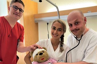 Drei Pflegekräfte versorgen auf einem Klinikbett spielerisch einen Teddybären mit Verband und Stethoskop