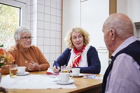 Eine Malteserin (M.) im Gespräch mit einer älteren Frau (l.) und einem älteren Herrn (r.) am Tisch sitzend. Foto: Malteser)