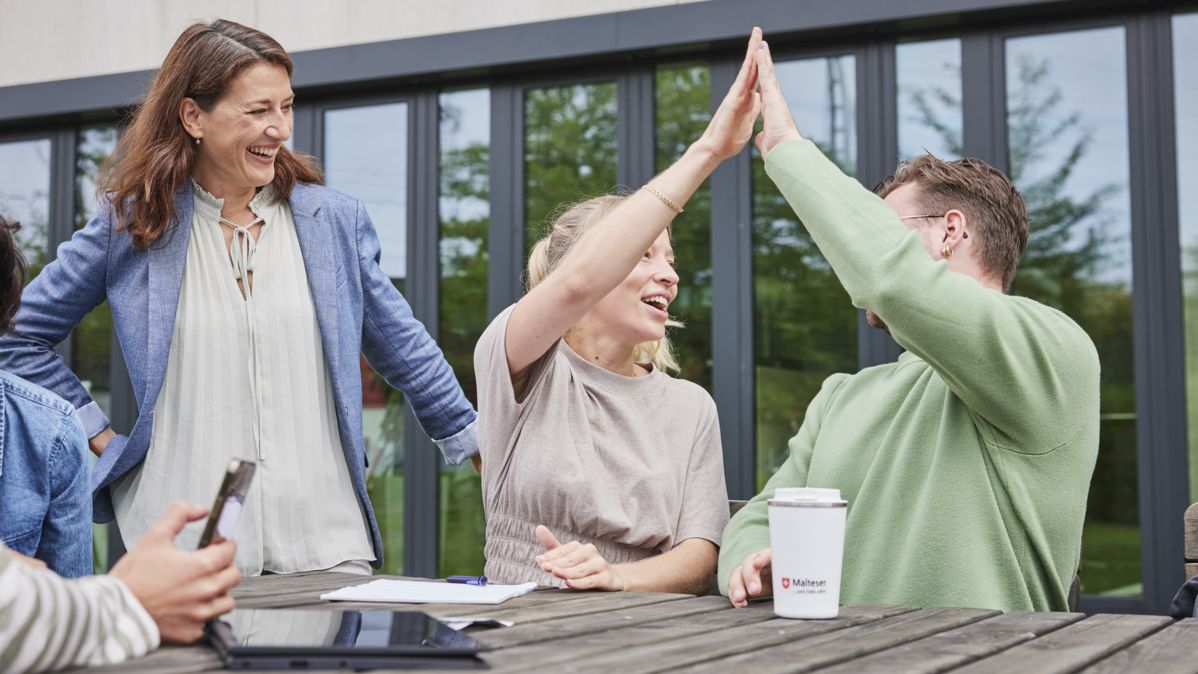 Zwei junge Menschen – links eine Frau, rechts ein Mann – sitzen an einem Outdoor-Holztisch vor einem Bürogebäude und klatschen erfreut jeweils mit einer Hand mittig oberhalb ihrer Köpfe ab. Ein weitere Frau steht links daneben und lacht mit den beiden.