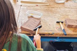 Eine Person sitzt an einer Werkbank und bearbeitet mit Werkzeugen eine Steinplatte. Auf dem Tisch liegen weitere Materialien und Arbeitsgeräte.