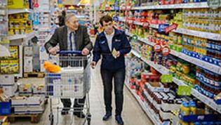 Ein älterer Herr geht mit einem Einkaufswagen, begleitet von einem Mann in Malteser Kleidung, zwischen Regalen eines Lebensmittelgeschäftes durch 