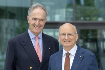 Zwei ältere Männer in Anzügen stehen nebeneinander im Freien vor einem modernen Gebäude.