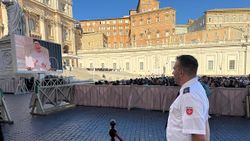 Ein Mann in Malteser Einsatzkleidung sieht auf einem Bildschirm Papst Leo, im Hintergrund Menschen auf dem Petersplatz in Rom. 