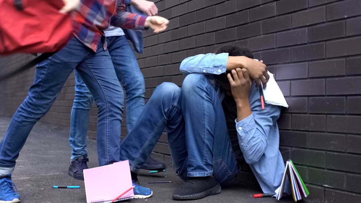 Ein Jugendlicher kauert an einer gemauerten Wand und hält schützend die Arme vor den Kopf; zwei weitere Personen stehen über ihm.