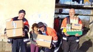 Drei Männer in Malteser Einsatzkleidung mit großen Paketen in den Händen stehen neben einem Kind, dem einer ein Paket überreicht, dahinter ein heruntergekommenes Gebäude