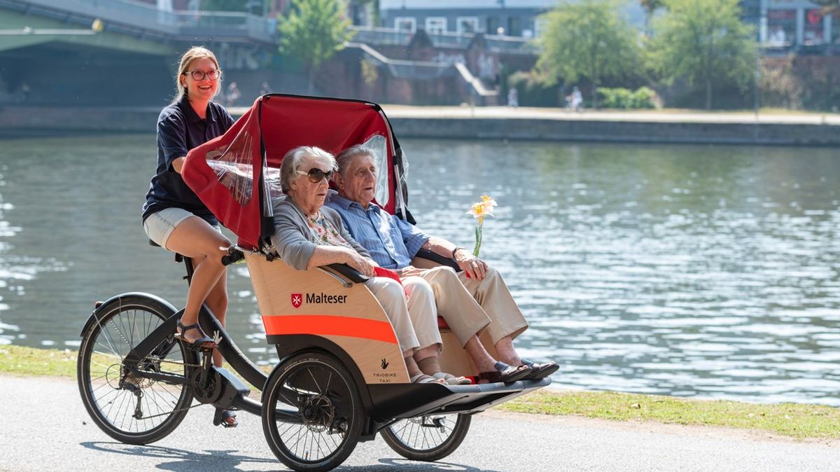 Älteres Paar sitzt in einer Rikscha, die von einer Frau gefahren wird, im Hintergrund ein Fluss.