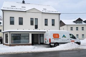 Ein helles zweistöckiges Gebäude mit verschneitem Dach und großem Schaufenster im Erdgeschoss. Vor dem Eingang steht ein weißer Transporter, der mit Aufklebern der Malteser und der Tafel beschriftet ist. Der Boden und die Umgebung sind von Schnee bedeckt.