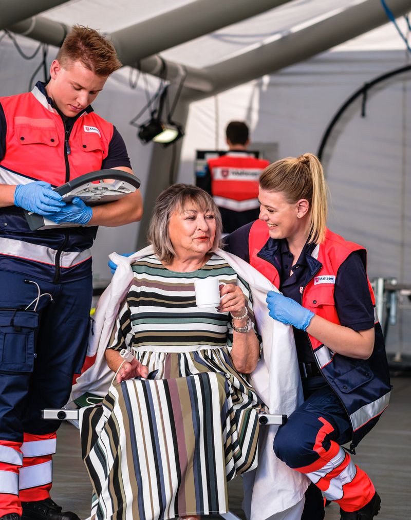 Zwei ehrenamtliche Einsatzkräfte betreuen in einem Zelt eine sitzende weibliche Person. Die Person hält eine Tasse in der Hand und bekommt gerade von einer weiblichen Einsatzkraft eine Decke übergelegt.