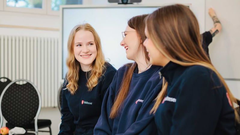 Drei junge, lächelnde Frauen mit Pullovern der Malteser in Deutschland unterhalten sich in einem Seminarraum. 