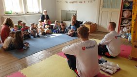 Eine Kindergruppe sitzt mit Erzieherinnen auf dem Fußboden. Zwei Sanitäter sprechen mit ihnen.