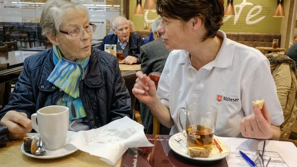 Zu sehen ist eine Frau und Malteser Mitarbeiterin, welche im Café sitzen.