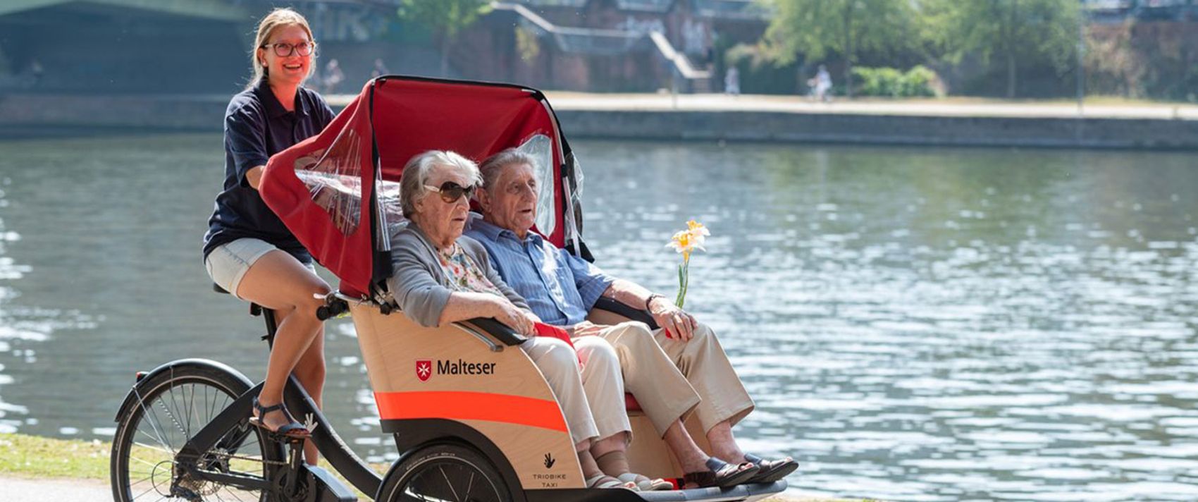Zwei Senioren sitzen in einer Malteser Rikscha und werden von einer Ehrenamtlichen an einem Flussufer entlanggefahren.