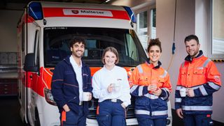 Vier Mitarbeitende des Malteser Rettungsdienstes stehen vor einem Rettungswagen.