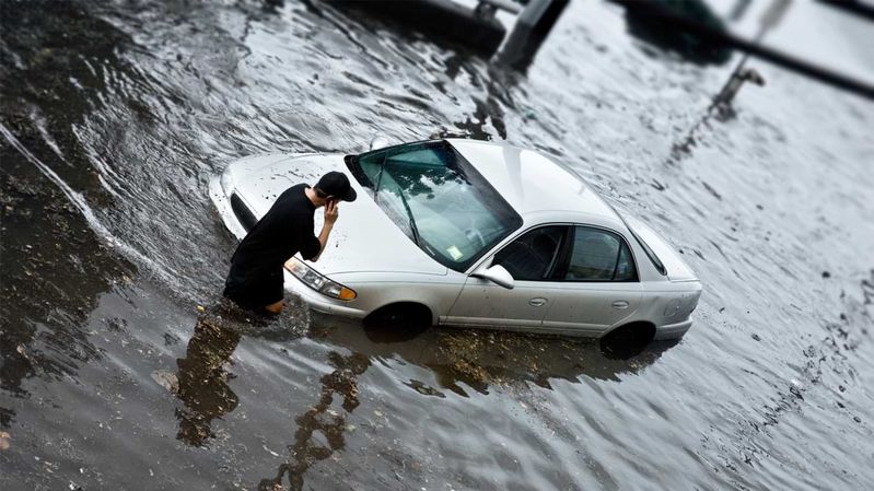 Ein junger Mann steht bei Hochwasser telefonierend vor einem Auto.