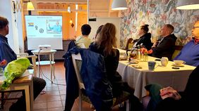 Menschen sitzen an einem Tisch. Sie schauen auf ein Bild auf einer Leinwand.