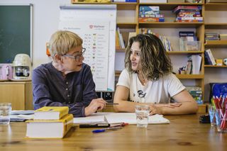 Zwei Frauen sitzen an einem Tisch vor einem Bücherregal und haben Bücher, Stifte und Zettel vor sich. Die linke Dame erklärt der rechten Dame etwas.