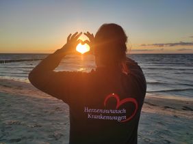 Eine Frau im Malteser Pulli steht mit dem Rücken zur Kamera, formt mit den Händen ein Herz.