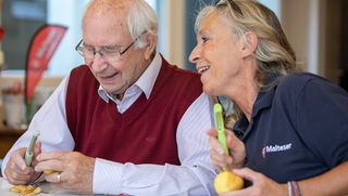 Ein älterer Mann und eine Mitarbeiterin der Malteser schälen gemeinsam Kartoffeln und lachen.