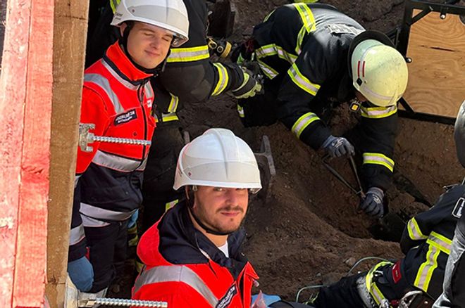 In einer Baugrube knien mehrere Einsatzkräfte mit Helmen und Schutzkleidung zwischen Erdreich und Baumaterial. Zwei Personen in roter Warnschutzkleidung befinden sich im Vordergrund, während weitere Einsatzkräfte in dunkler Schutzkleidung an einer Rettungs- oder Bergungsmaßnahme arbeiten.