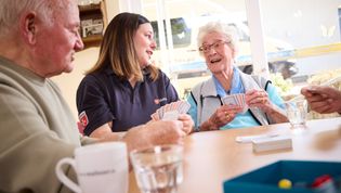 Mehrere Personen sitzen gemeinsam an einem Tisch und spielen Karten. Auf dem Tisch stehen Gläser und eine Tasse. Im Hintergrund fällt durch ein großes Fenster Tageslicht in den Raum, draußen ist ein Fahrzeug der Malteser zu erkennen.