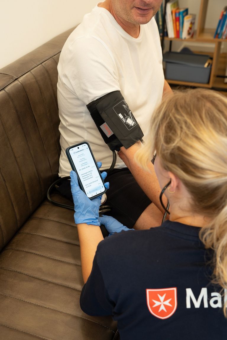 Das Bild zeigt eine Malteser-Mitarbeiterin, die einem Patienten den Blutdruck misst und die Daten in den TI-Messenger eingibt. 