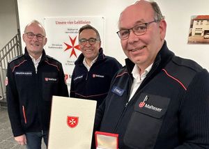 Drei Männer in Malteser Dienstbekleidung. Der Vordere hält Urkunde und Medaille.
